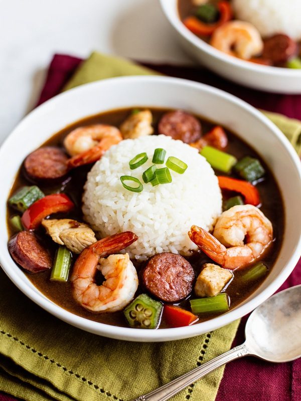 A bowl of dark brown gumbo with shrimp, sliced andouille sausage, chicken, okra, celery and red bell pepper around a mound of white rice topped with green onions.
