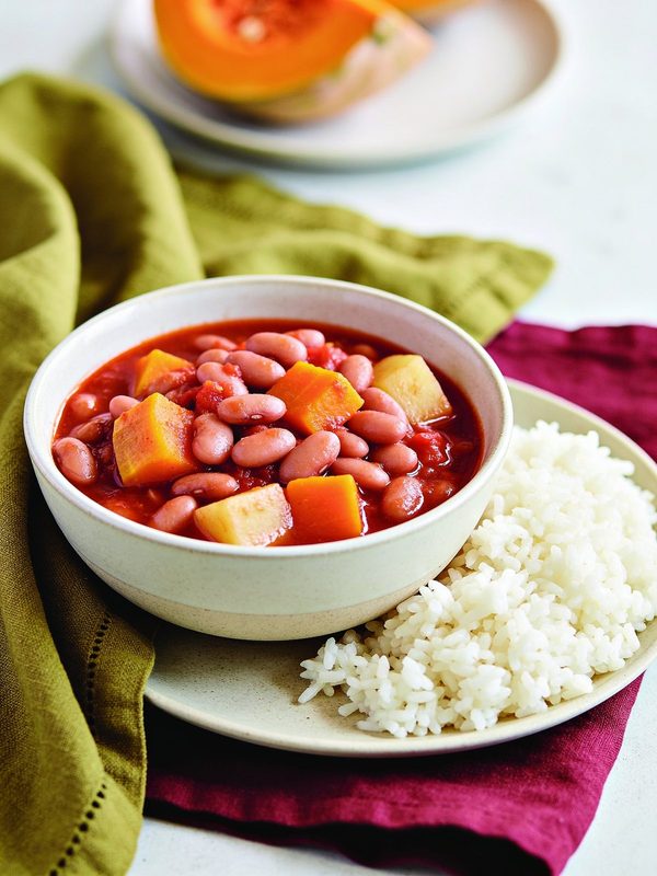 A bowl of stewed pink beans with cubes of calabaza and potato in tomato sauce, next to white rice.