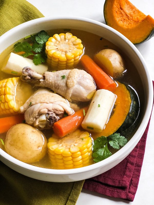 A bowl of clear chicken soup with drumsticks, corn on the cob, yuca, potatoes, carrots, pumpkin and cilantro.