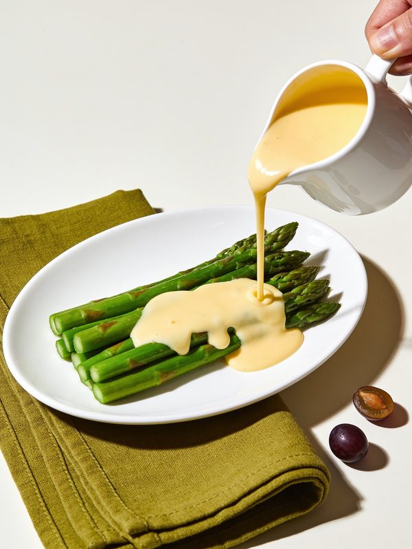 A small white pitcher of pale yellow hollandaise sauce poured over steamed green asparagus.