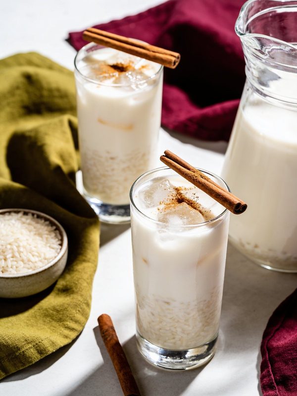 Two glasses of creamy horchata over ice with cinnamon sticks, next to a glass pitcher and a small bowl of rice.