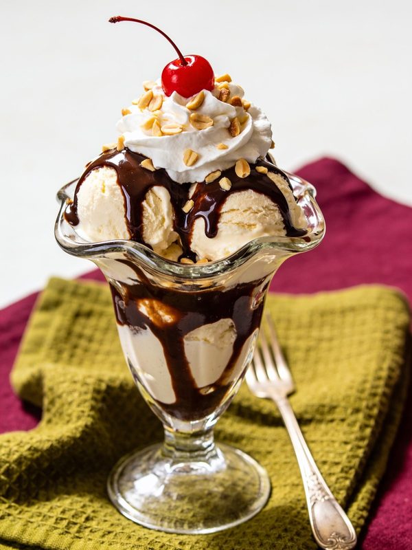A hot fudge sundae in a tall glass with vanilla ice cream, chocolate sauce, whipped cream, chopped peanuts and a cherry.