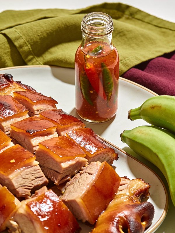 Chunks of roast pork with glossy amber skin on a platter, with a bottle of pique and green plantains beside them.