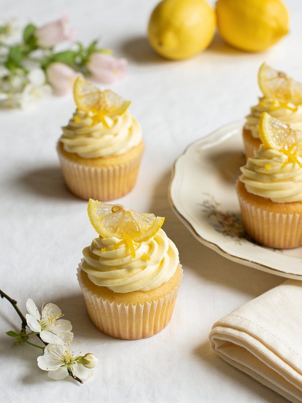 Lemon cupcakes with pale yellow buttercream swirls topped with candied lemon slices and lemon zest, with whole lemons in the background.