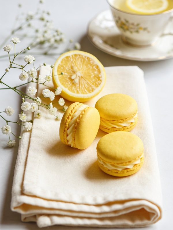 Bright yellow macarons with piped lemon cream on a cream linen napkin, with a halved lemon, white flowers and a cup of tea.