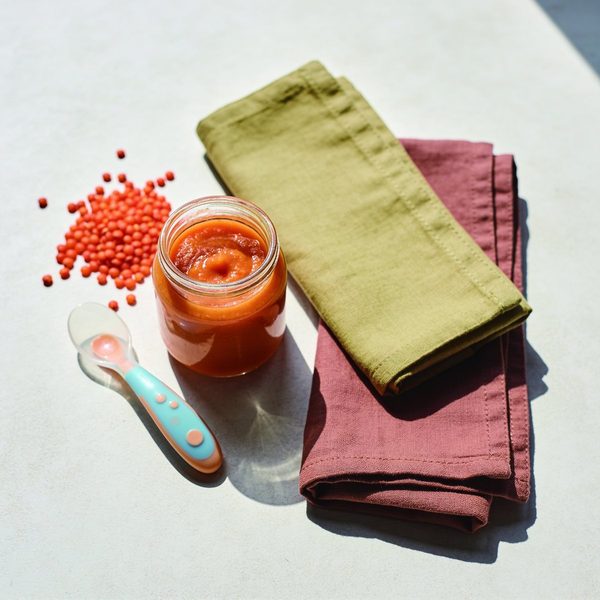 A small glass jar of smooth orange red lentil puree with a baby spoon and a little pile of red lentils beside it.