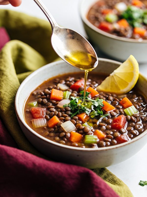 A bowl of brown lentil soup with diced carrots, celery, onion and tomato, a drizzle of olive oil being spooned on top, parsley and a lemon wedge.