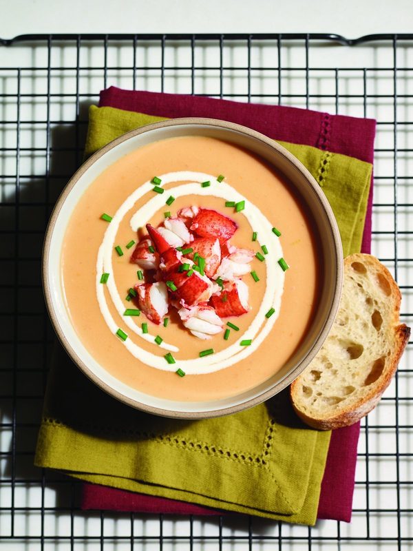 A bowl of smooth pale coral lobster bisque with a swirl of cream, chunks of red and white lobster meat and chopped chives, with a slice of crusty bread.