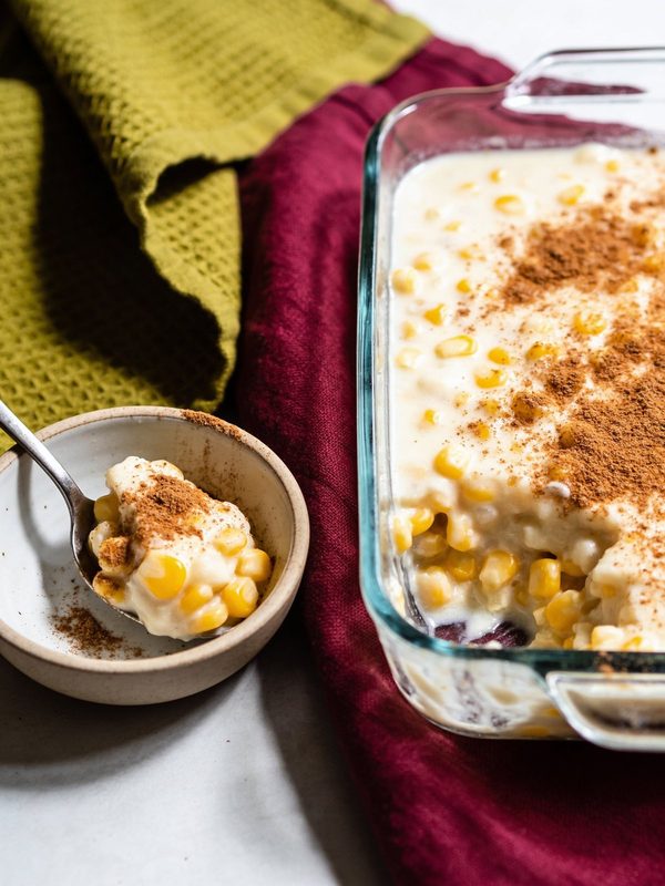 A glass baking dish of creamy corn and coconut pudding with whole kernels and cinnamon on top, next to a small bowl with a spoonful.