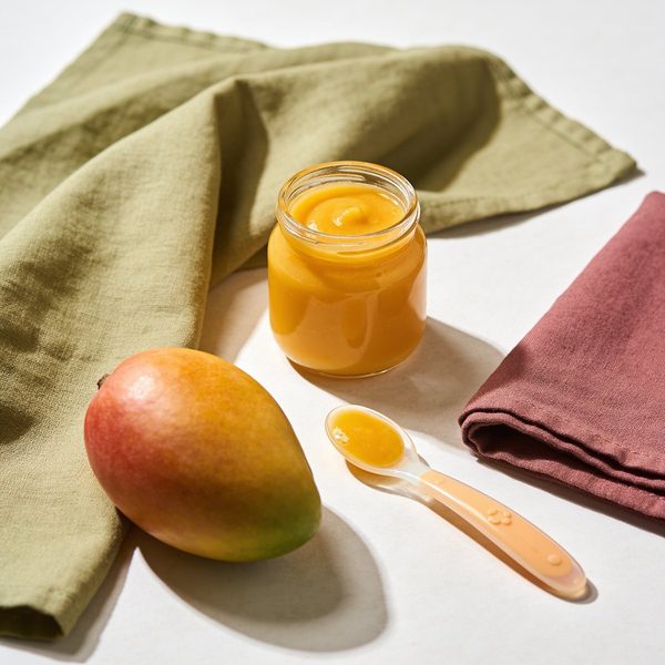 A small glass jar of smooth bright yellow mango puree with a silicone baby spoon and a whole ripe mango beside it.