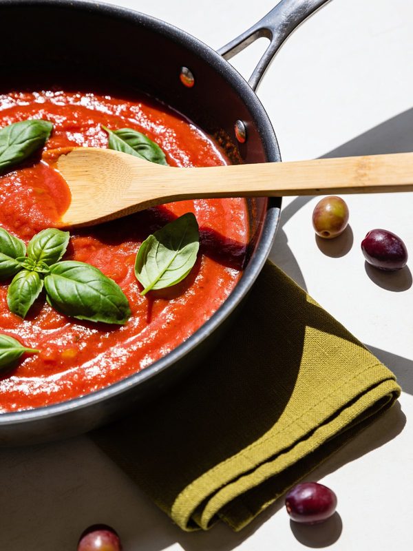 A pot of bright red marinara sauce with fresh basil leaves and a wooden spoon.