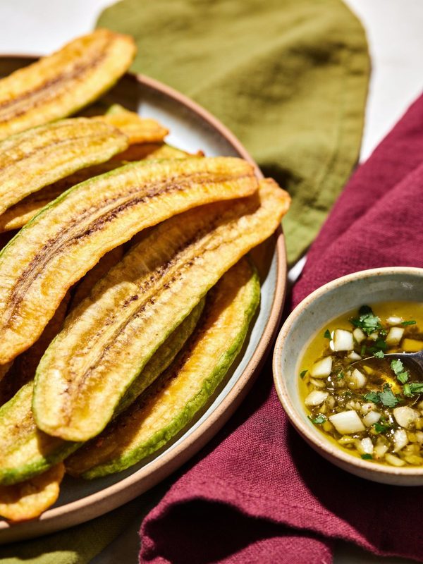Long, thin golden green plantain chips piled on a plate, with a small bowl of garlic mojo with olive oil and parsley.