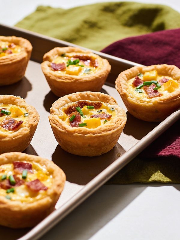 Mini quiches in golden pastry cups filled with bacon, cheddar and chives on a baking sheet.