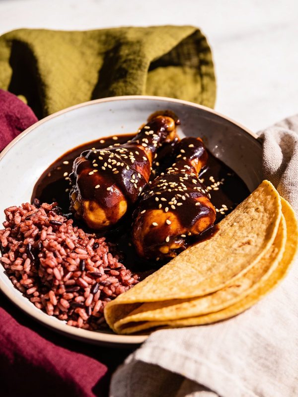 Two chicken drumsticks covered in dark mole poblano and sesame seeds in a bowl with red rice and folded corn tortillas.