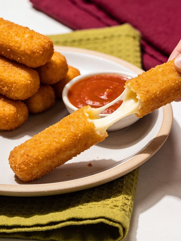 Golden breaded mozzarella sticks on a plate, one pulled apart to show a long stretch of melted cheese, with a small bowl of marinara sauce.