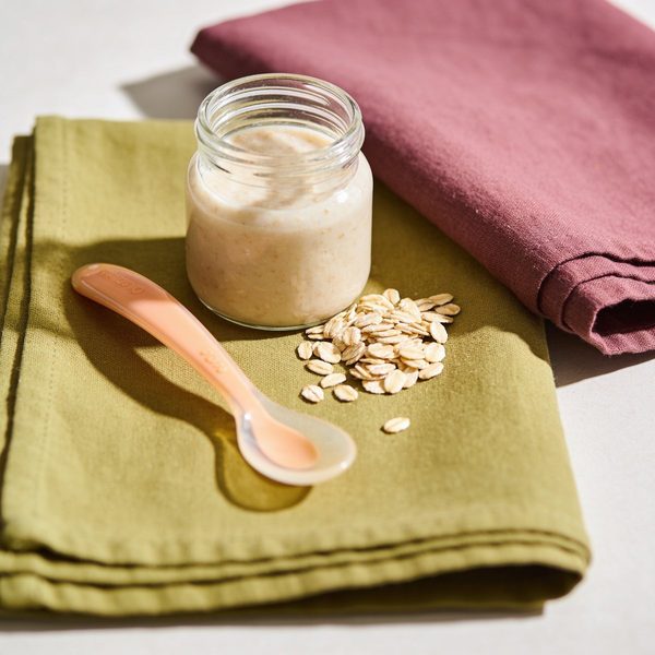 A small glass jar of smooth, creamy oatmeal baby cereal with a silicone baby spoon and a little pile of rolled oats beside it.