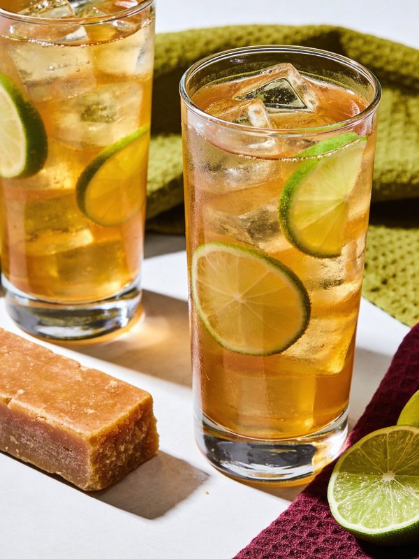 Two tall glasses of amber papelón con limón with ice and lime slices, next to a block of papelón and a halved lime.