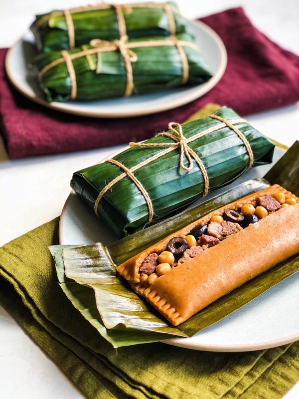 Pasteles wrapped in banana leaves and tied with string, one unwrapped to show orange masa with pork, olives and chickpeas.