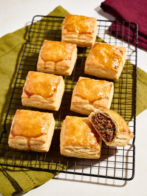 Square golden puff pastries with a shiny glaze on a cooling rack, one cut open to show the ground beef filling.