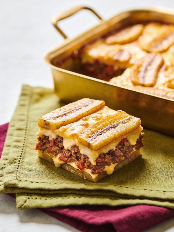 A square of pastelón with layers of fried sweet plantain, beef picadillo and melted cheese, in front of the baking pan.