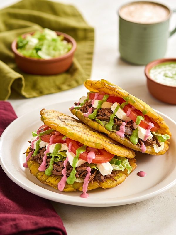 Two patacones of crispy flattened green plantain filled with shredded beef, lettuce, tomato and white cheese, drizzled with green and pink sauces.