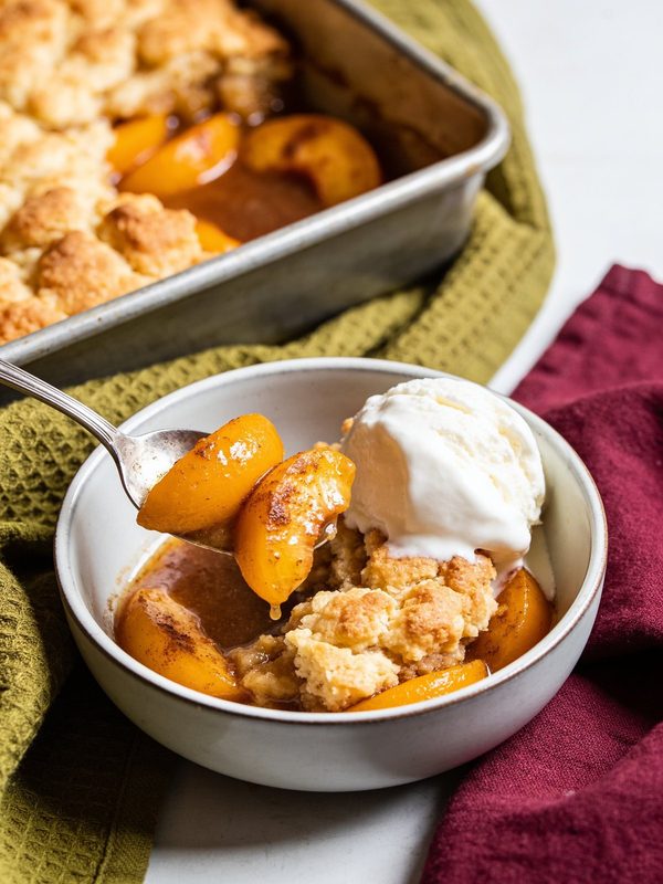 A bowl of peach cobbler with golden peach slices, biscuit topping and a scoop of vanilla ice cream, with the baking pan behind.