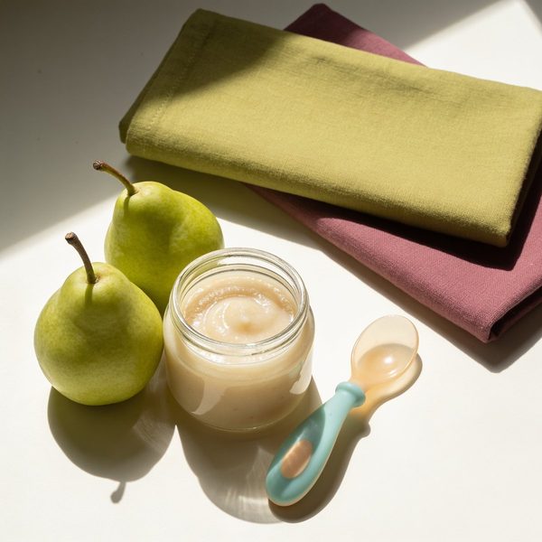 A small glass jar of smooth, creamy pear puree with a silicone baby spoon and two fresh green pears beside it.