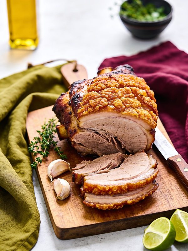 Roast pernil with golden crackling skin, partly sliced on a wooden board with garlic, thyme and lime wedges.