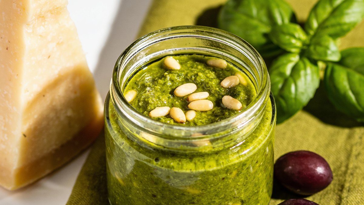 A small glass jar of bright green basil pesto with pine nuts, a wedge of parmesan and fresh basil leaves.