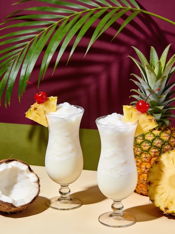 Two frosty piña coladas in tall glasses garnished with pineapple wedges and cherries.