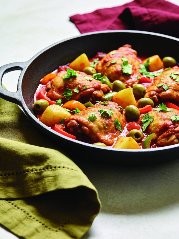 Golden chicken thighs in tomato sauce with potatoes, green olives, red peppers and cilantro in a cast iron skillet.