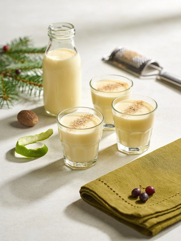 Small glasses and a clear glass bottle of creamy pale yellow Venezuelan ponche crema dusted with grated nutmeg, with Christmas pine sprigs around.