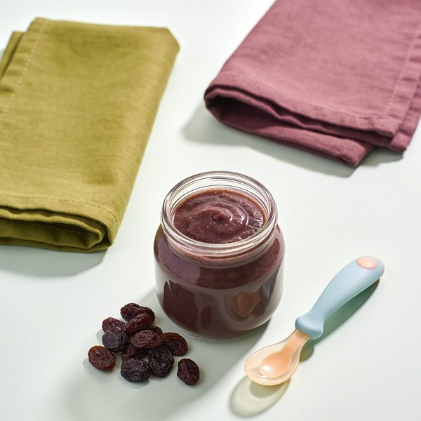 A small glass jar of smooth, dark prune puree with a silicone baby spoon and whole dark plums beside it.