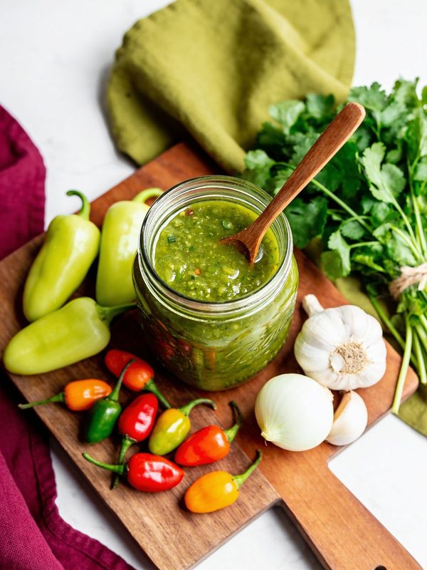 A glass jar and an ice cube tray filled with bright green Puerto Rican sofrito, with ají dulce and culantro beside them.