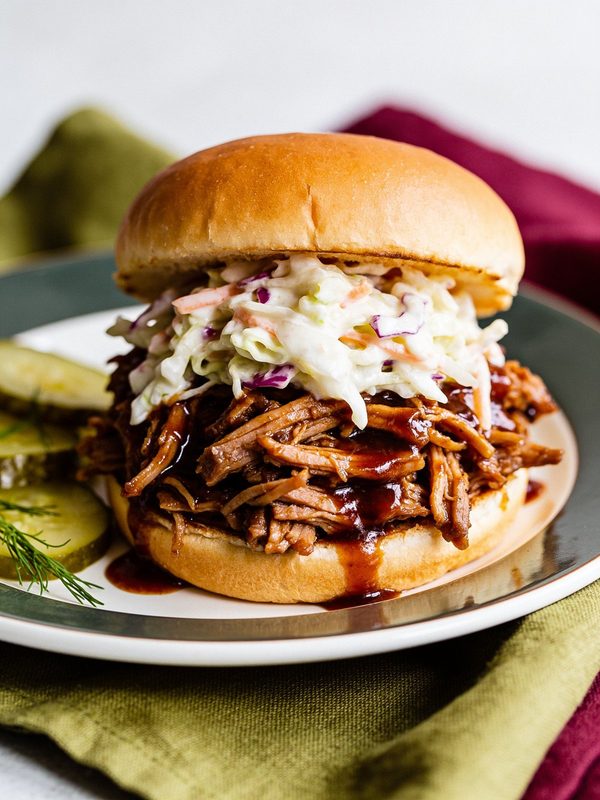 A soft bun piled with shredded barbecue pork and creamy coleslaw, with dill pickle slices on the plate.