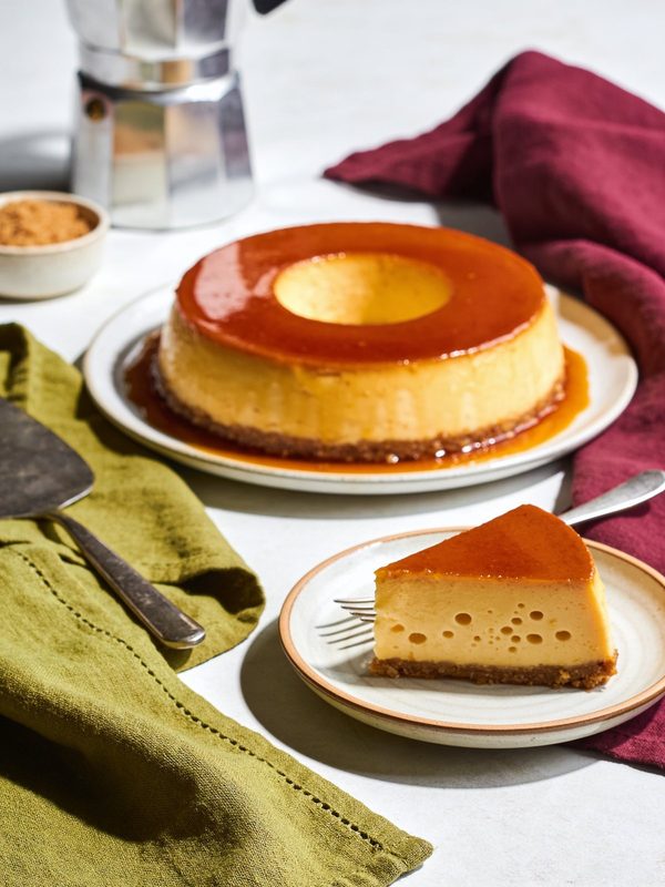 A ring-shaped quesillo glazed with amber caramel, with a slice on a plate showing its tiny holes.