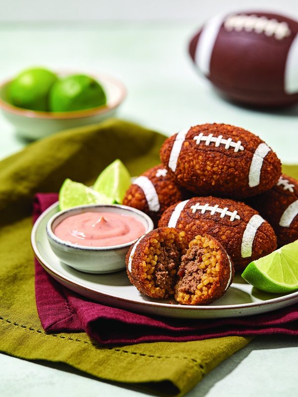 Fried bulgur quipes decorated as little footballs, one cut open to show the beef filling, with lime wedges and mayo-ketchup.