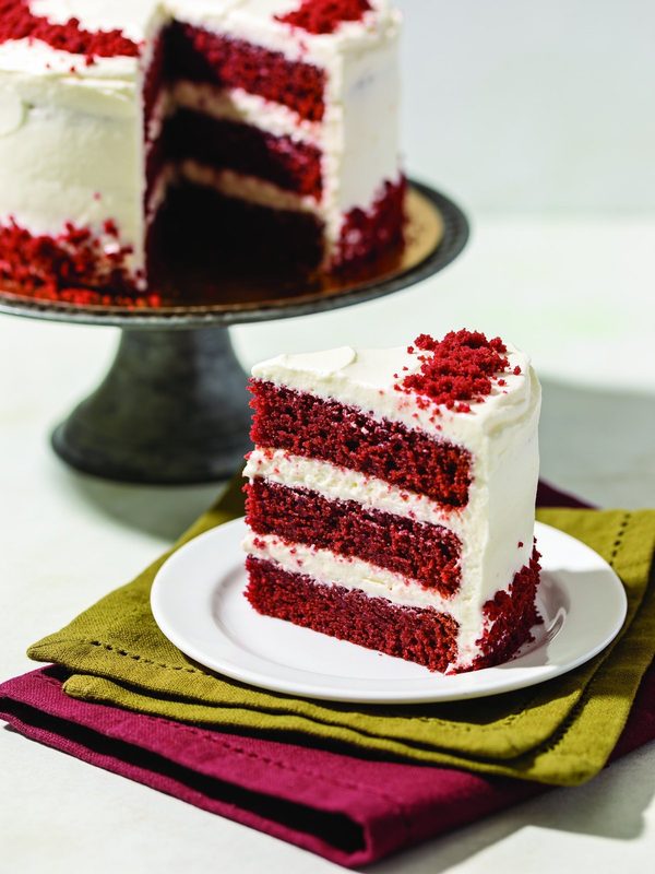 A slice of red velvet cake with three red layers and white cream cheese frosting, topped with red crumbs, beside the cake on a stand.