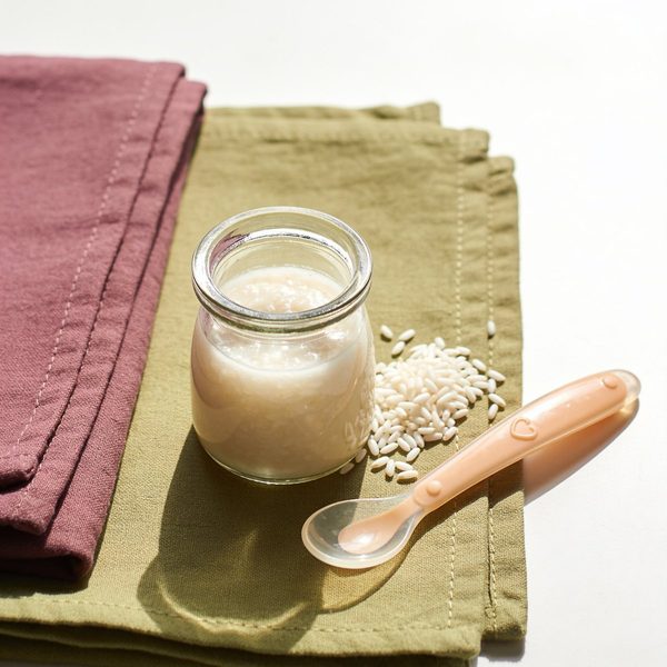 A small glass jar of creamy white rice baby cereal with a silicone baby spoon and uncooked rice grains beside it.