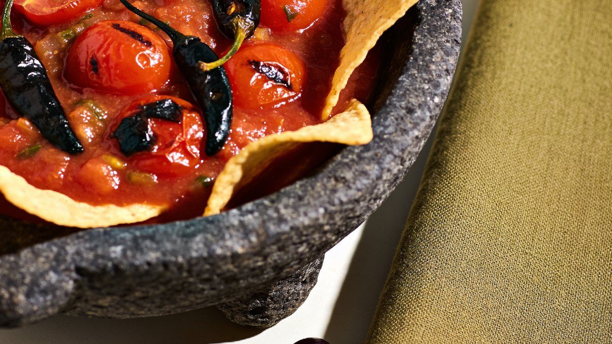 A stone bowl of red roasted tomato salsa with charred tomatoes, chiles and tortilla chips around it.