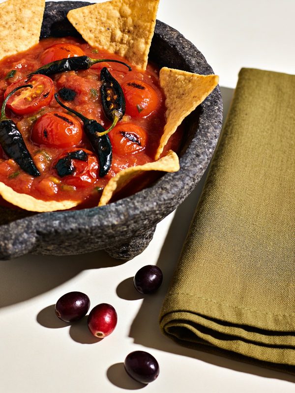 A stone bowl of red roasted tomato salsa with charred tomatoes, chiles and tortilla chips around it.