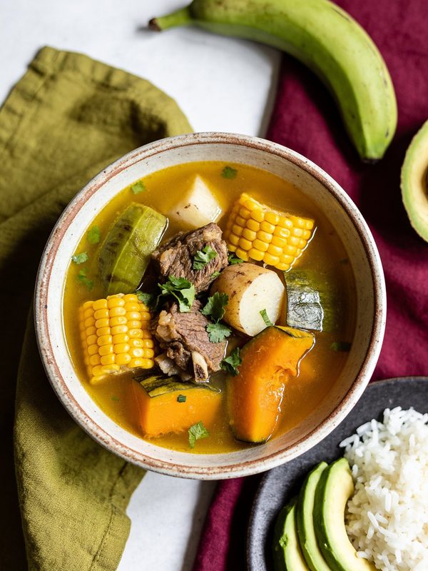 A bowl of sancocho with beef, corn on the cob, calabaza, potato and plantain in a golden broth, with avocado and rice on the side.