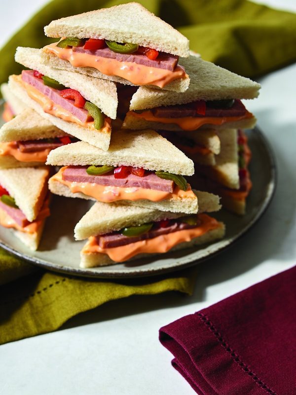 Crustless white bread triangles filled with an orange cheese spread, luncheon meat and pimientos, stacked on a plate.