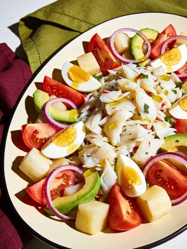 A platter of flaked salt cod with yuca, tomato wedges, avocado, halved boiled eggs and red onion rings, drizzled with olive oil.
