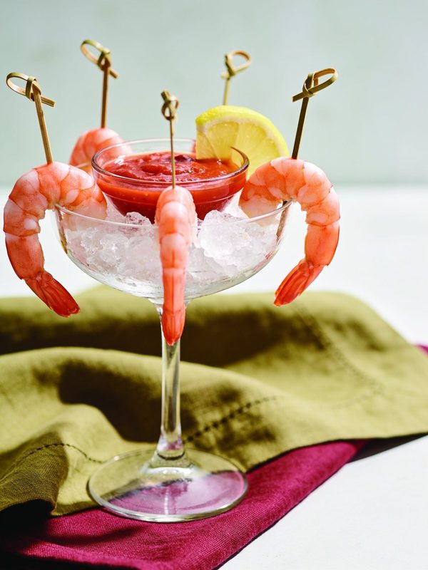 Chilled pink shrimp on bamboo picks hanging from a glass coupe of crushed ice with red cocktail sauce and a lemon wedge.