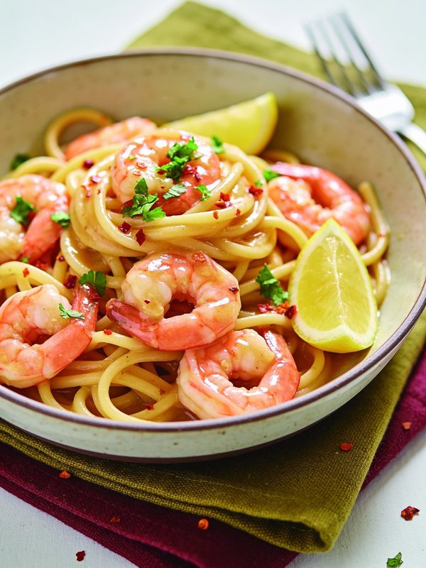 Linguine with pink shrimp in a garlic butter and lemon sauce, topped with parsley and red pepper flakes, with lemon wedges in a wide bowl.