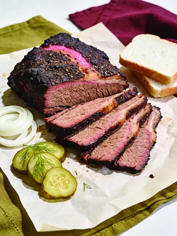 Slices of brisket with a dark peppery crust and a pink ring fanned out on butcher paper, with dill pickles, sliced white onion and white bread.