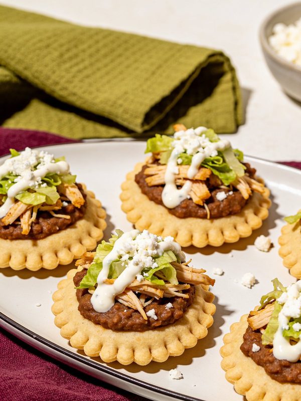 Round corn sopes with pinched edges on a platter, topped with refried beans, shredded chicken, lettuce, crumbled queso fresco and crema.