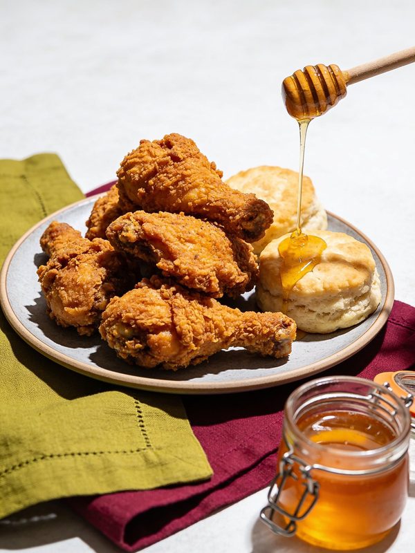 A plate of golden, crunchy fried chicken drumsticks and thighs with buttermilk biscuits and honey being drizzled from a wooden dipper.
