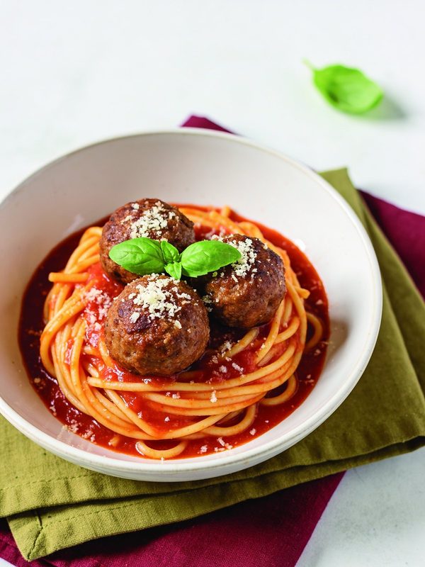 A shallow white bowl of spaghetti in red marinara sauce with three browned meatballs, grated Parmesan and a sprig of fresh basil.